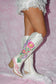 White cowgirl boots with pink and purple hand-painted flowers and a pink bow, displayed on pink lace fabric.