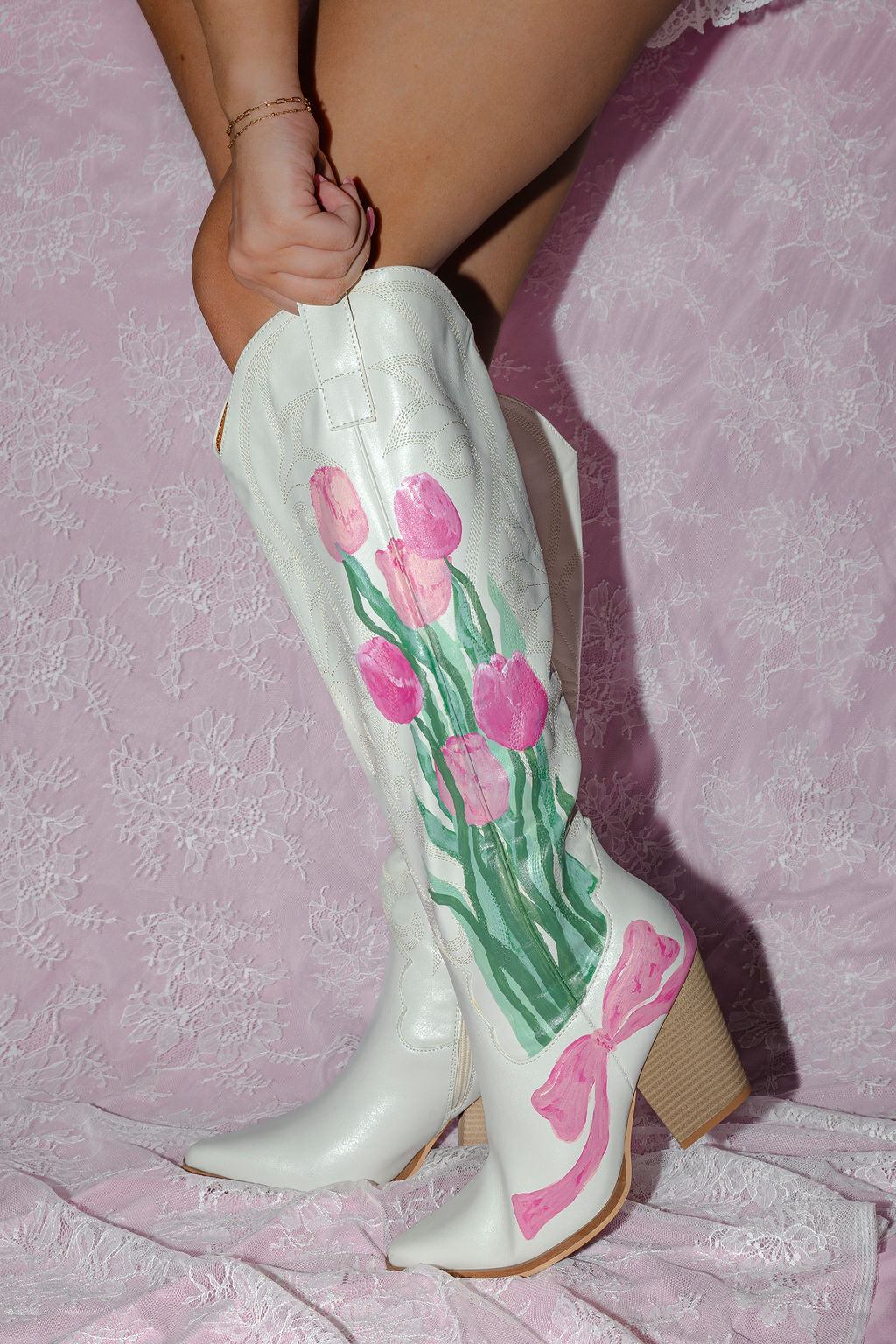 Hand-painted white cowgirl boots with pink tulips and pink bow styled on pink lace background